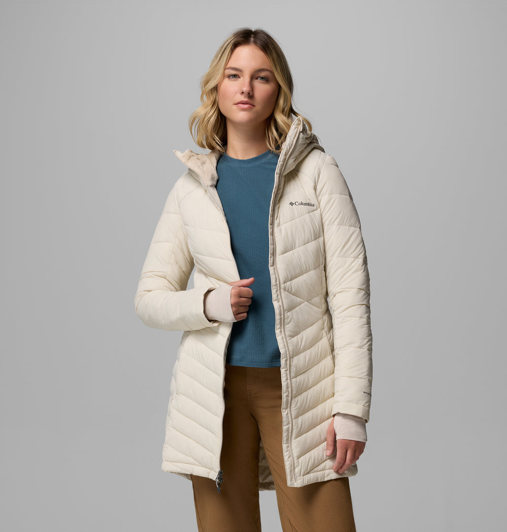 אישה הלובשת את מעיל החורף "Joy Peak II Mid Jacket" של קולומביה IL — מעיל חורף מרופד בצבע בז' בהיר עם ברדס — עומדת בביטחון, לבושה בחולצה כחולה ומכנסיים בצבע חום בהיר, על רקע אפור.