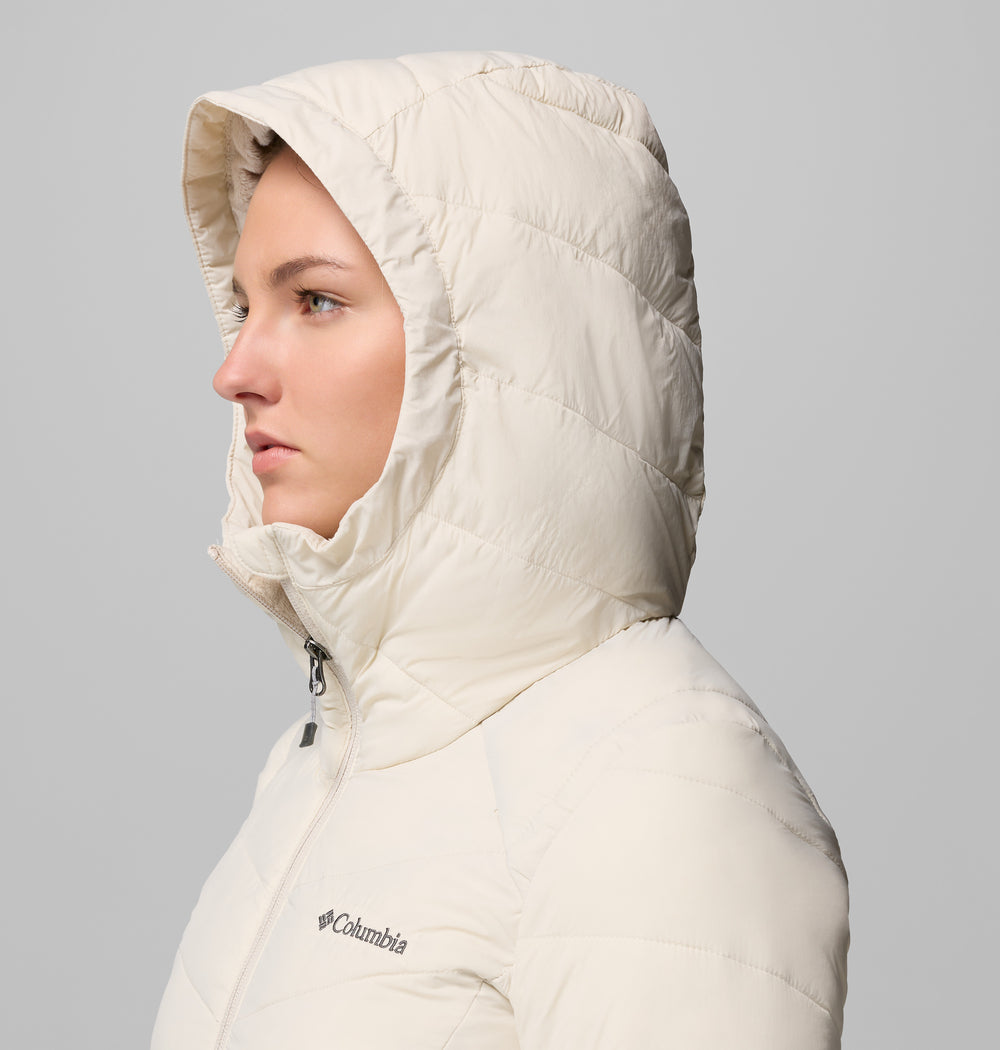 אדם הנראה בפרופיל לובש מעיל פוך בהיר מדגם Joy Peak II Mid Jacket של המותג ColumbiaIL, כשהכובע מורם, מכסה את רוב ראשו וממסגר את פניו, על רקע אפור אחיד.