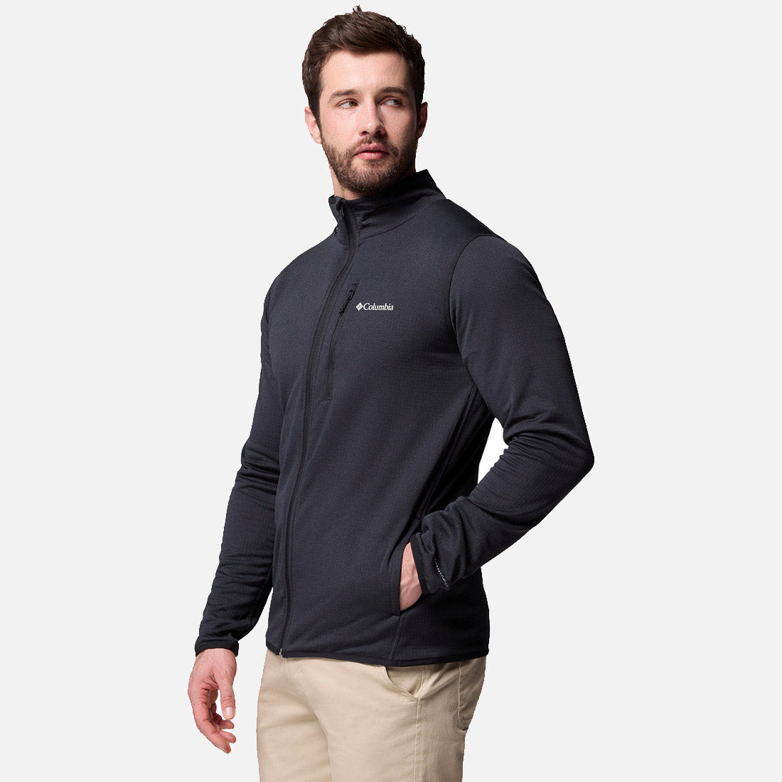גבר לבוש בחולצת קולומביה ישראל Essential Hike Grid Full Zip M בצבע אפור כהה ובמכנסיים בצבע בז', עומד כשידו האחת בכיס, מביט לצד על רקע אפור בהיר אחיד.