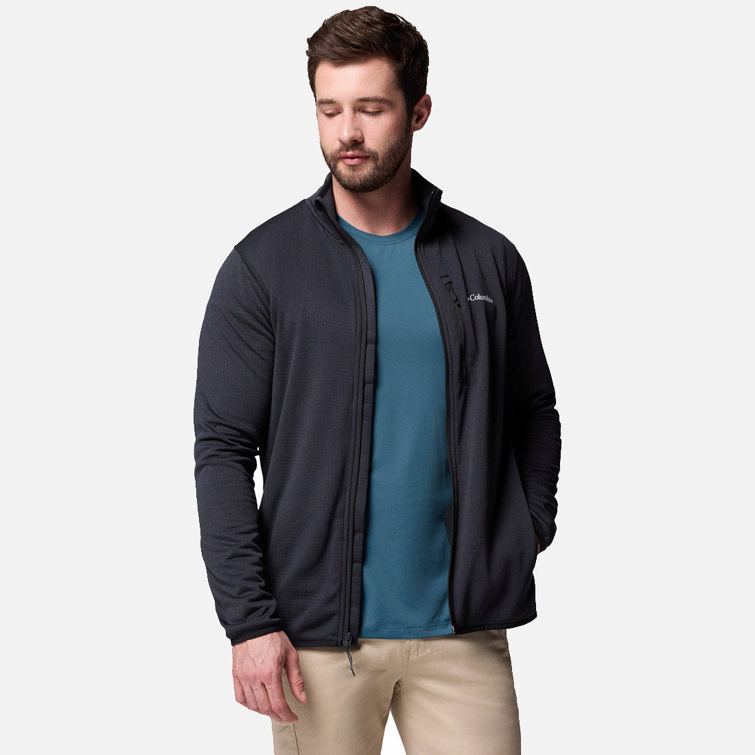 גבר עם שיער חום קצר וזקן לובש את מעיל ה-Essential Hike Grid Full Zip M של קולומביה ישראל מעל חולצה בצבע טורקיז ומכנסיים בצבע בז', עומד עם יד אחת בכיס על רקע לבן חלק.