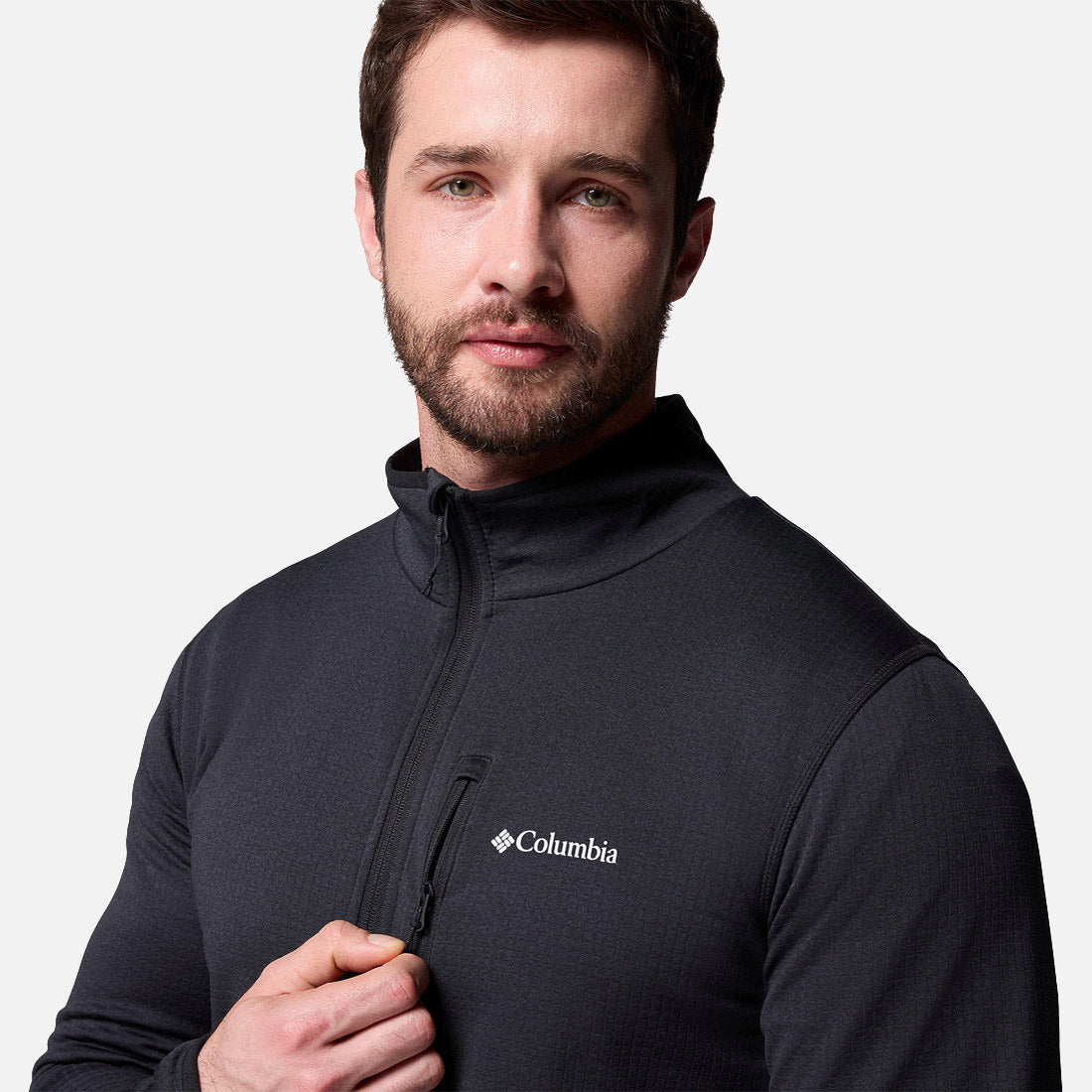 גבר מזוקן, לבוש ב-Essential Hike Grid Full Zip M של קולומביה ישראל בצבע שחור, עומד על רקע לבן חלק, מביט למצלמה ואוחז ברוכסן ליד חזהו.